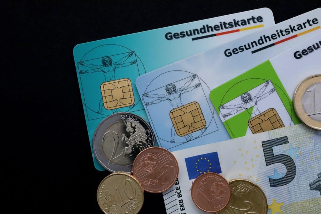 Als Beamte in die private oder gesetzliche Krankenversicherung? Kosten in Form von Gesundheitskarten mit Münzen und Geldschein